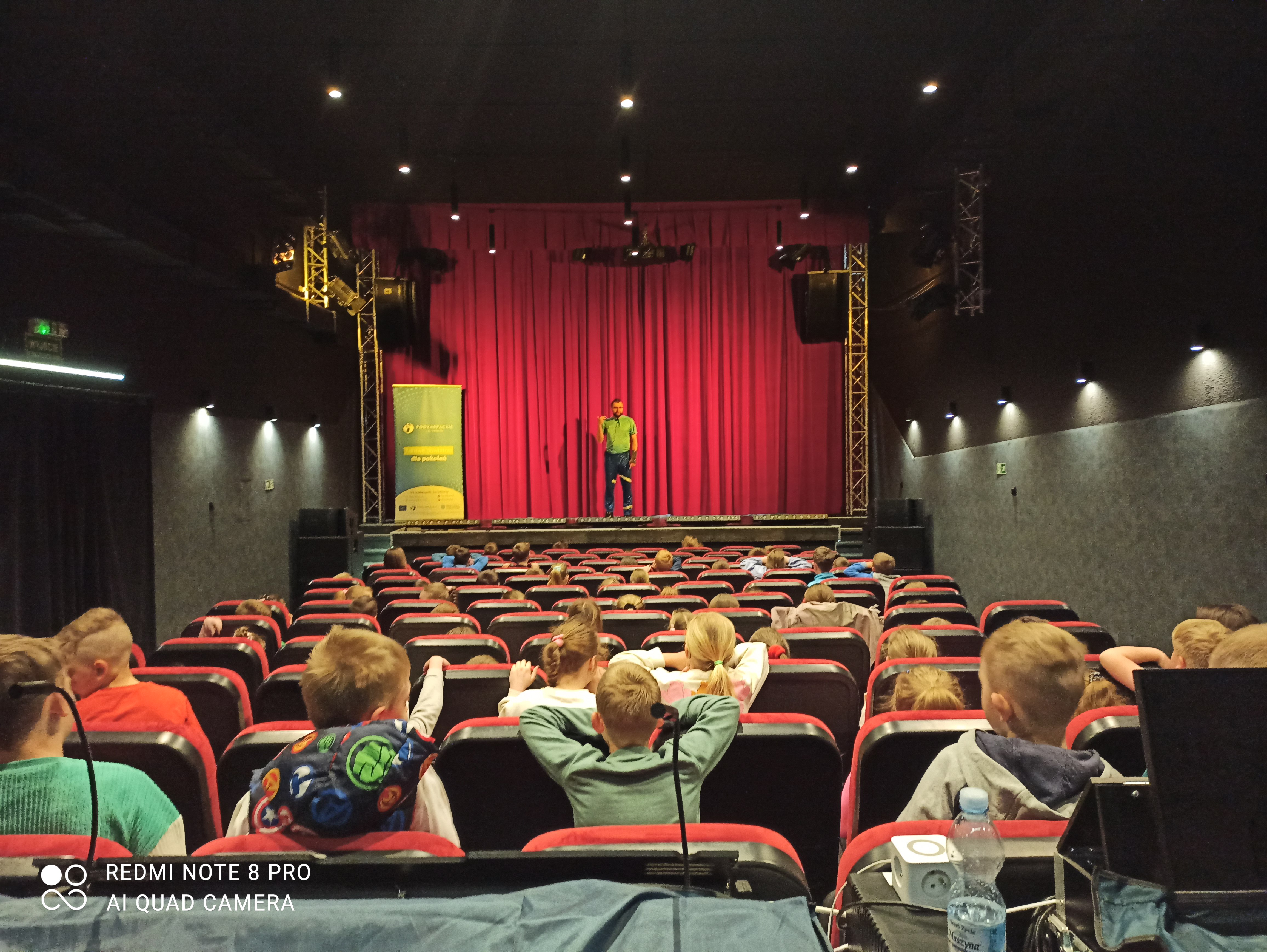 Sala kinowa wypełniona dziećmi, na scenie stoi prelegent przed czerwoną kurtyną. Widok z perspektywy widza.